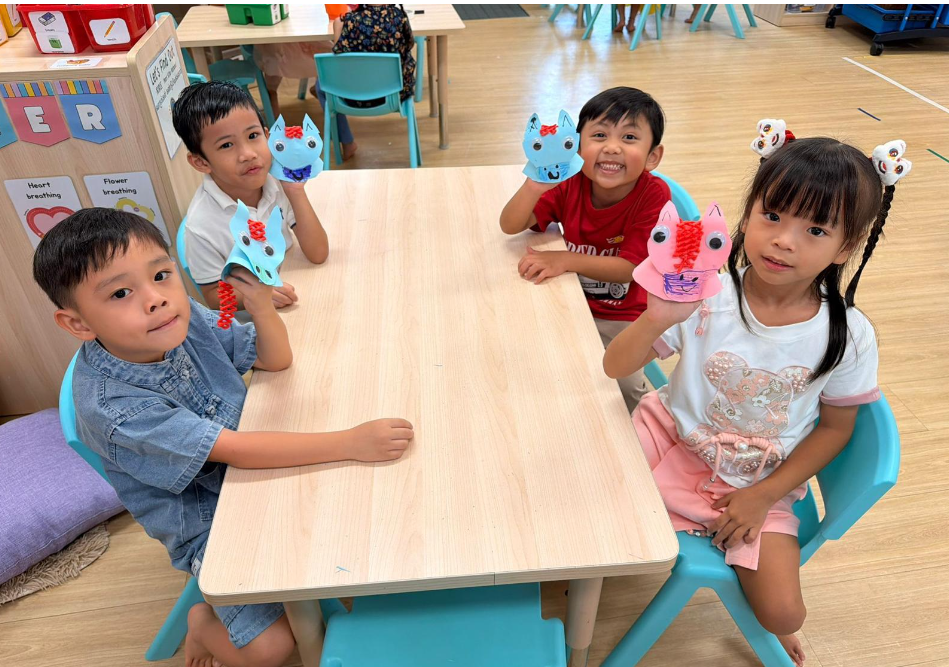 Four children at a classroom table hold colorful paper animal puppets and smile at the camera.